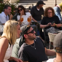 Menschen sitzen im Sommer in einem Biergarten und schauen lachend nach vorn.