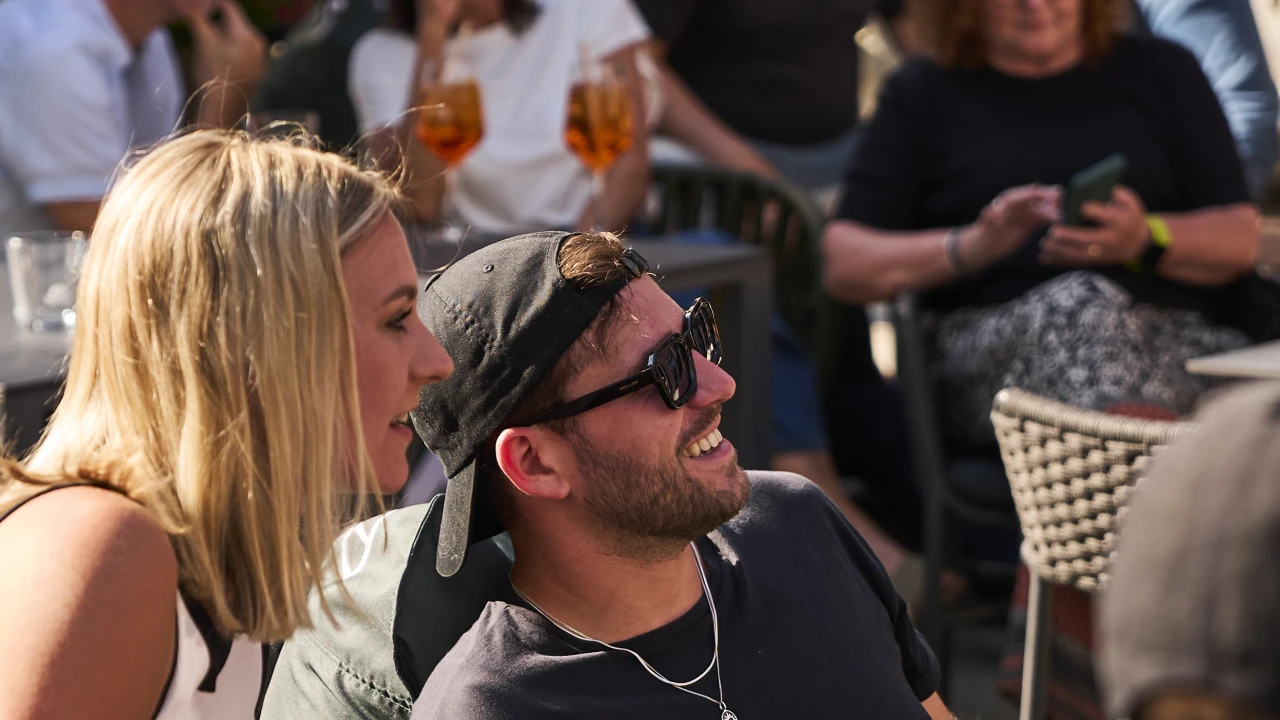 Menschen sitzen im Sommer in einem Biergarten und schauen lachend nach vorn.