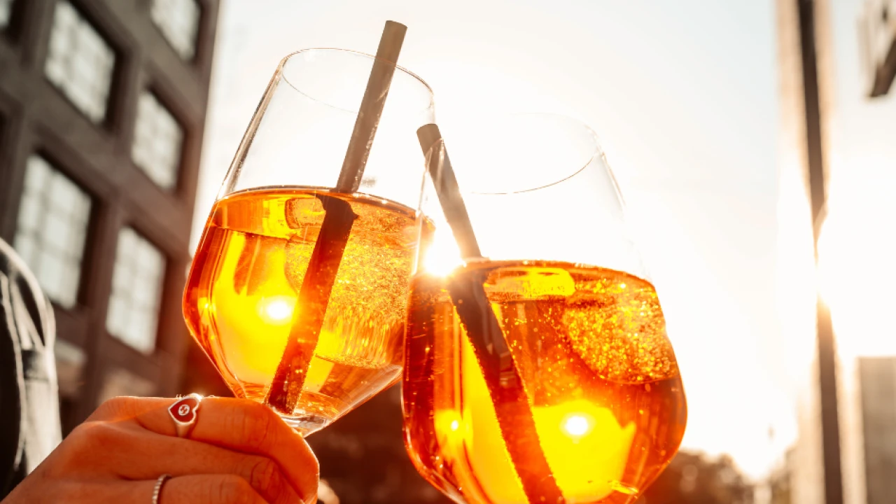 Bild von zwei Aperol Spritz Getränken in der Sonne. Im Hintergrund zwei Gebäude der Macherei München