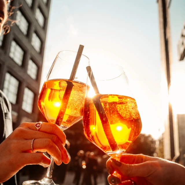 Bild von zwei Aperol Spritz Getränken in der Sonne. Im Hintergrund zwei Gebäude der Macherei München